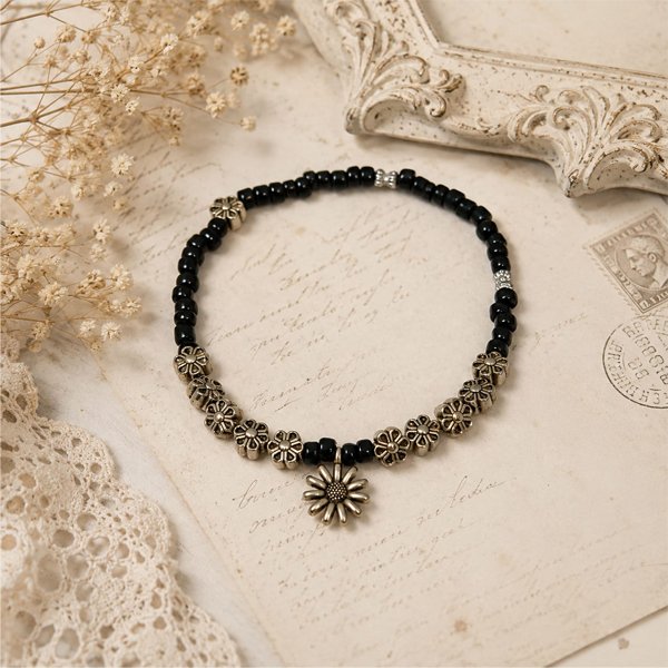 Bloom-&-Beads-Anklet Handmade black beaded anklet with antique silver floral beads and a daisy charm, displayed on a soft vintage cream background.
