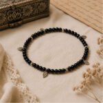 Handcrafted Forest Whisper Anklet with black glass beads, antique silver leaf charms, and spacer beads on a soft beige fabric background.