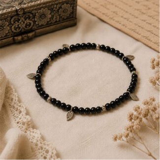 Handcrafted Forest Whisper Anklet with black glass beads, antique silver leaf charms, and spacer beads on a soft beige fabric background.
