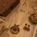 Srida Peacock Royale Necklace Set featuring a layered pearl necklace with an antique gold-tone peacock pendant in red and green enamel, paired with matching jhumka earrings.