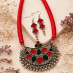Srida Raga Red Necklace Set featuring a handcrafted oxidized silver-tone crescent pendant with red and green enamel detailing, braided red cotton thread, matching hook earrings, and decorative ghungroo beads.
