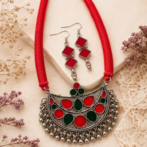 Srida-Raga-Red-Necklace-Set Srida Raga Red Necklace Set featuring a handcrafted oxidized silver-tone crescent pendant with red and green enamel detailing, braided red cotton thread, matching hook earrings, and decorative ghungroo beads.