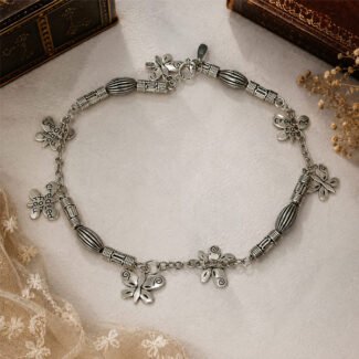 Whispering Wings Anklet in oxidised silver finish featuring butterfly charms and engraved floral charms, handcrafted with textured metal beads, photographed on a light vintage background.