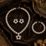 White Pearl Bloom Necklace Set featuring a medium-length pearl necklace with a five-pearl flower centerpiece, matching pearl bracelet with lobster clasp, and hook earrings displayed on an elegant antique background.White Pearl Bloom Necklace Set featuring a medium-length pearl necklace with a five-pearl flower centerpiece, matching pearl bracelet with lobster clasp, and hook earrings displayed on an elegant antique background.