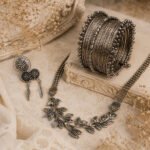 Handcrafted oxidized silver-tone necklace with leaf-inspired centerpiece, matching feather earrings, statement ring, and stacked bangles displayed on a vintage cream background.