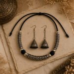 Handcrafted Srida Antique Aura Choker Set featuring an oxidized silver finish, black adjustable braided dori, and matching oxidized hook jhumka earrings on a vintage antique background.
